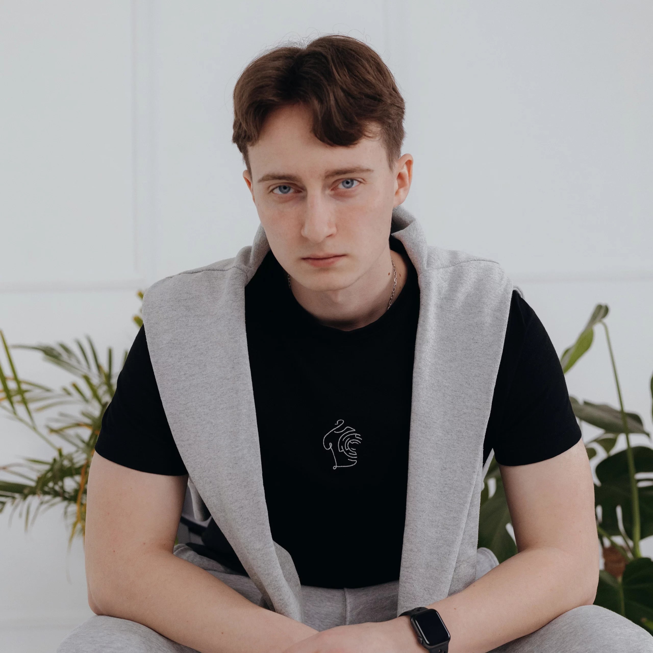 Mężczyzna siedzący w jasnym wnętrzu, mający na sobie oryginalny black minimaze t-shirt z delikatnym, białym haftem oraz szarą bluzę zarzuconą na ramiona.