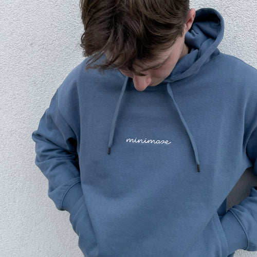 Mężczyzna z głową skierowaną w dół, prezentujący niebieską bluzę z kapturem oraz białym napisem na piersi, czyli oryginalny blue minimaze hoodie.