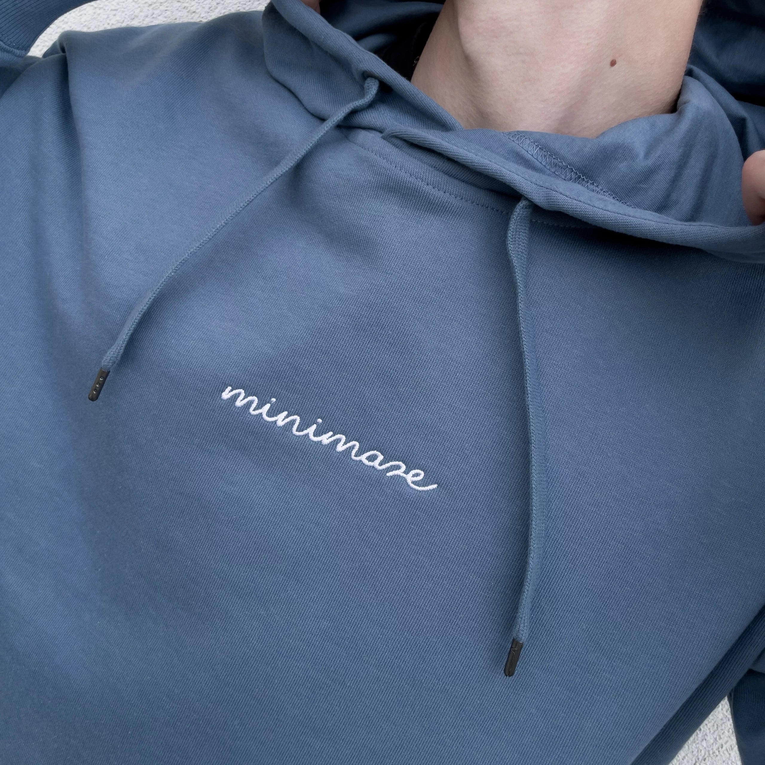 Kadrowany widok na klatkę piersiową w niebieskiej bluzie z kapturem, prezentujący biały, odręczny napis oryginalnego blue minimaze hoodie.
