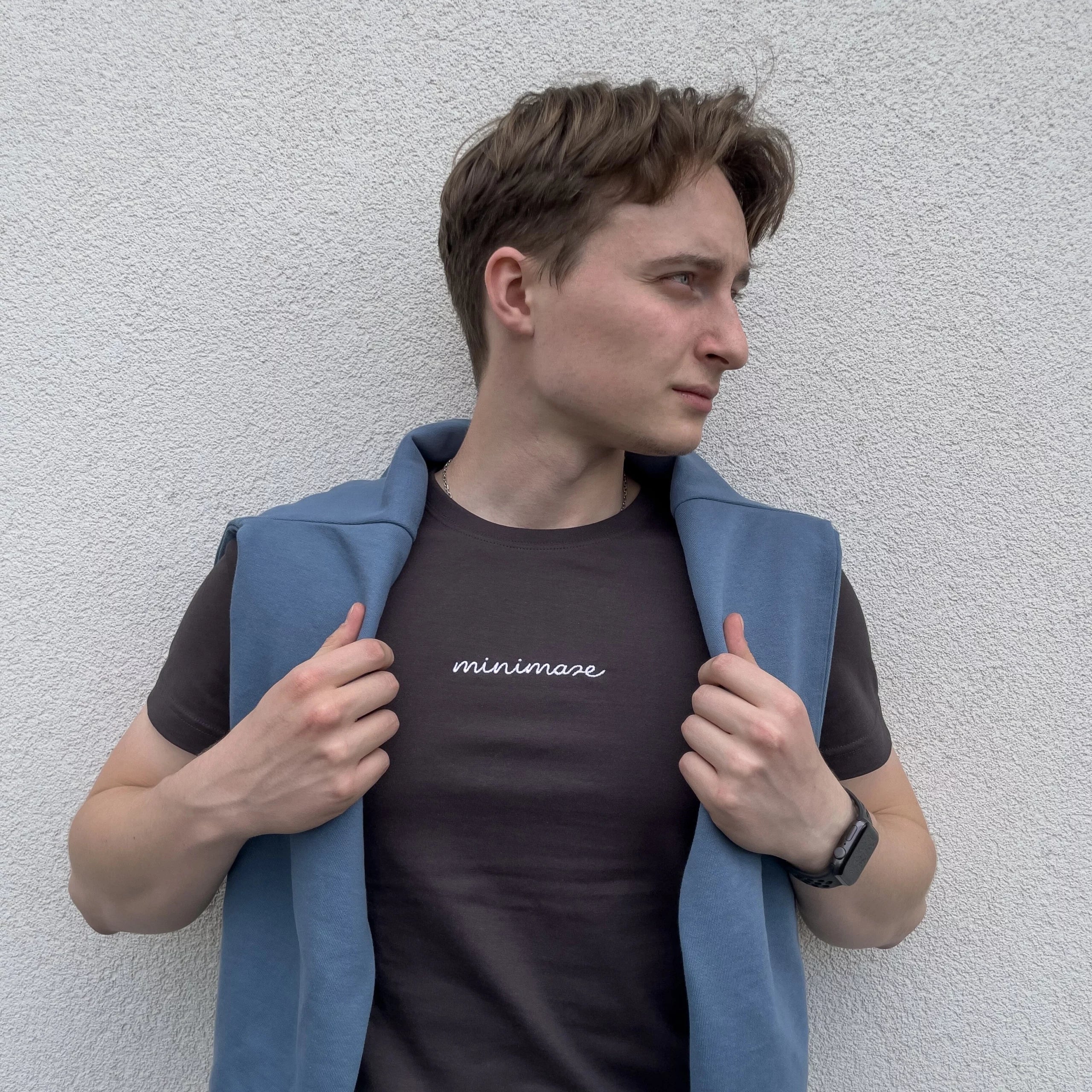 Profil mężczyzny w niebieskiej bluzie zarzuconej na ramiona, prezentującego grafitową koszulkę z białym napisem na piersi, czyli oryginalny graphite minimaze t-shirt.