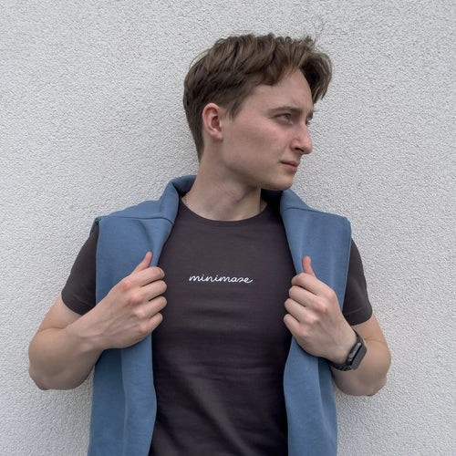 Profil mężczyzny w niebieskiej bluzie zarzuconej na ramiona, prezentującego grafitową koszulkę z białym napisem na piersi, czyli oryginalny graphite minimaze t-shirt.