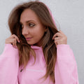 Profil młodej kobiety trzymającej dłońmi kaptur różowej bluzy z delikatnym, białym napisem na piersi, prezentujący stylowy pink minimaze hoodie.