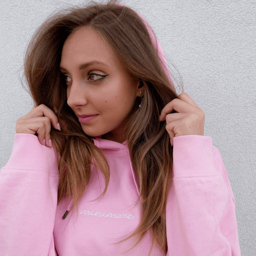 Profil młodej kobiety trzymającej dłońmi kaptur różowej bluzy z delikatnym, białym napisem na piersi, prezentujący stylowy pink minimaze hoodie.