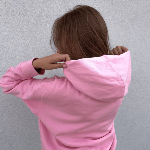 Widok od tyłu na kobietę zakładającą kaptur różowej bluzy dresowej na tle jasnej ściany, prezentujący tył i krój oryginalnego pink minimaze hoodie.