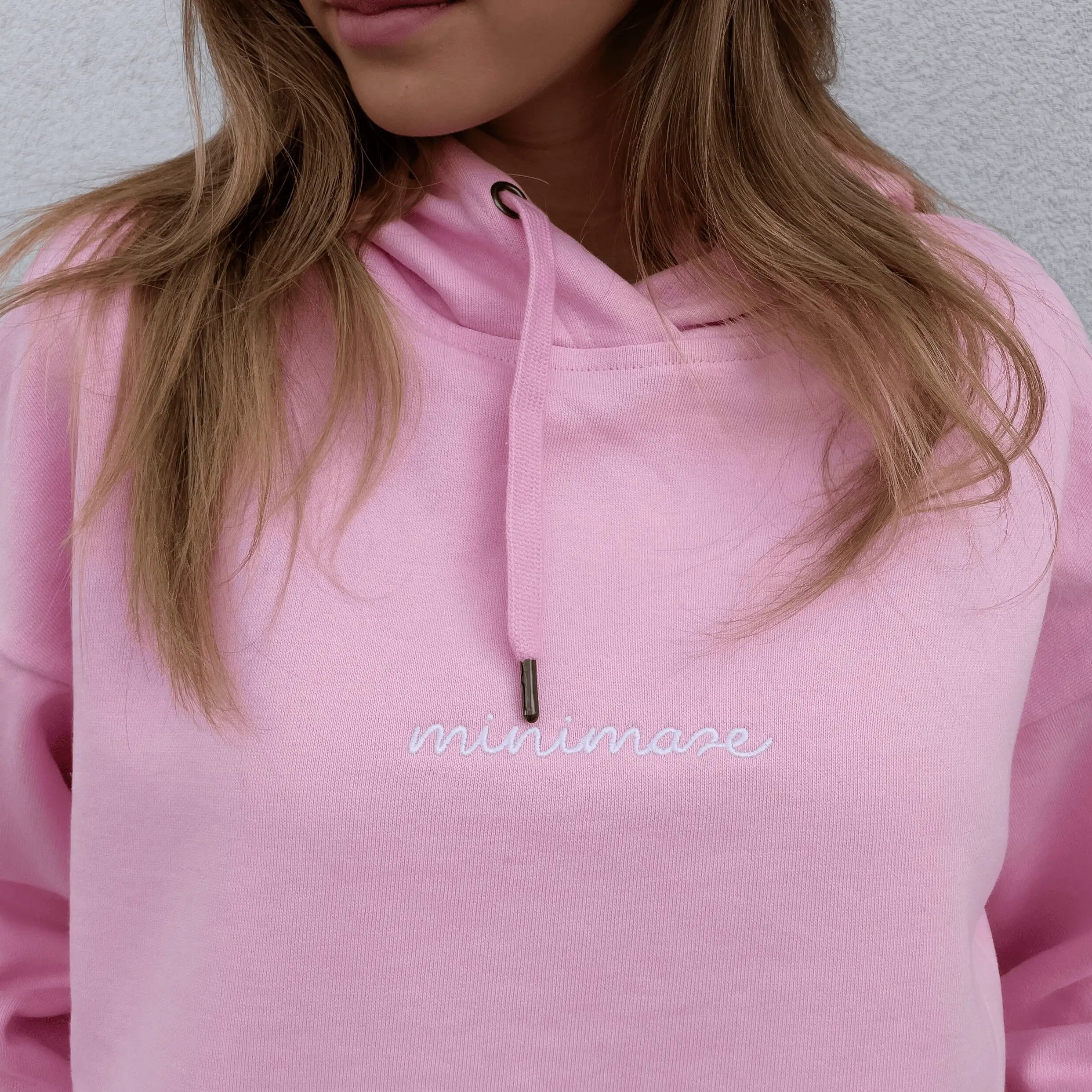 Kadrowany widok na klatkę piersiową w różowej bluzie z kapturem, prezentujący biały, odręczny napis oryginalnego pink minimaze hoodie.