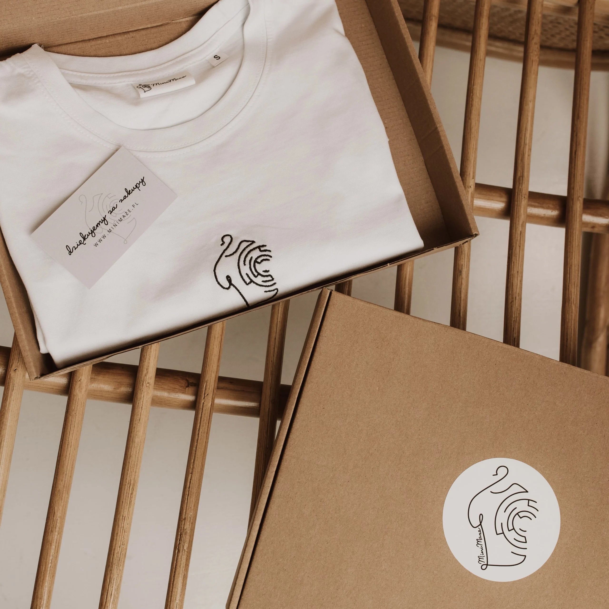 Biały t-shirt z minimalistycznym haftem zapakowany w ekologiczne pudełko z logo, prezentujący oryginalny white minimaze t-shirt na bambusowym krześle.