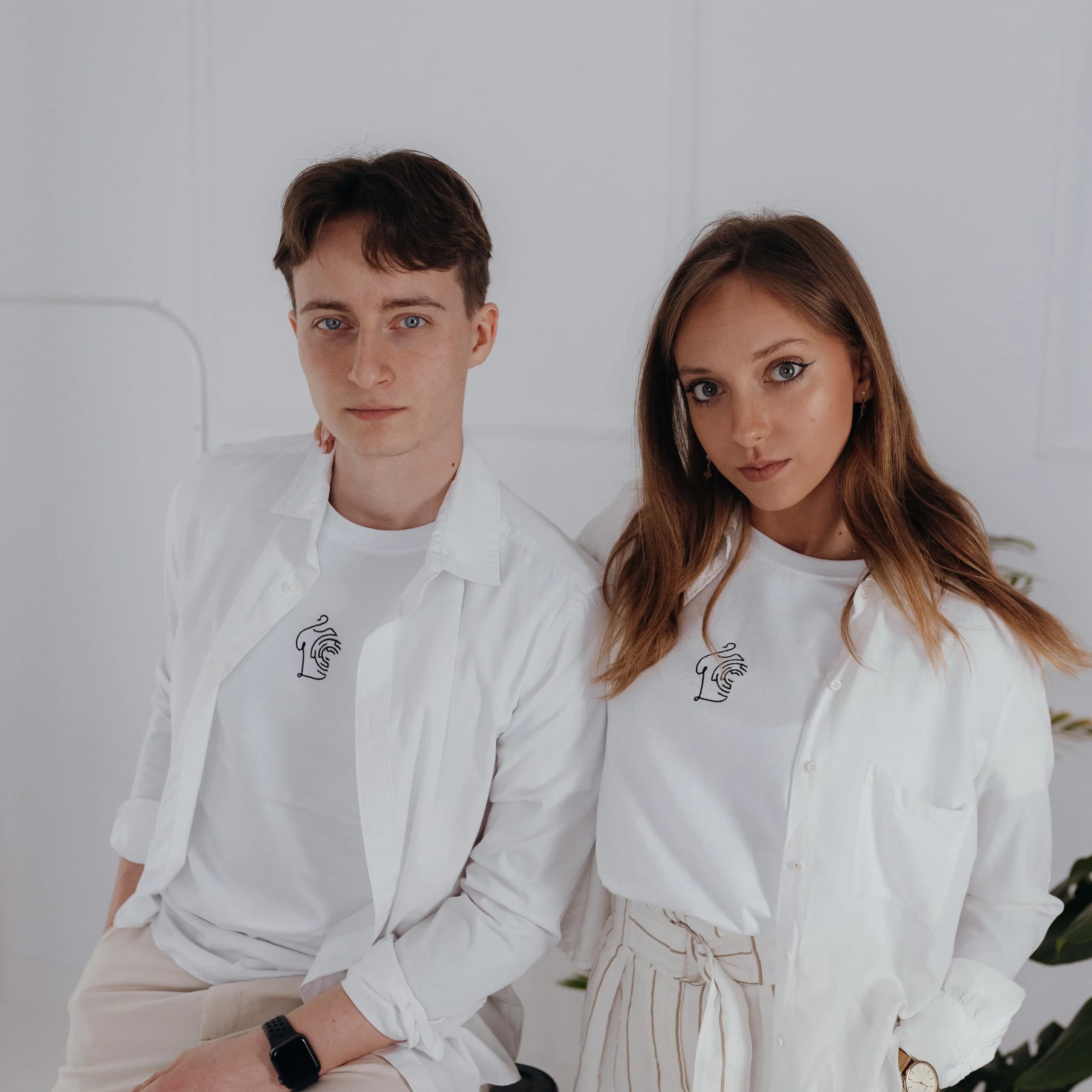 Kobieta i mężczyzna w białych rozpiętych koszulach prezentują dopasowany white minimaze t-shirt z minimalistycznym, czarnym haftem na piersi.