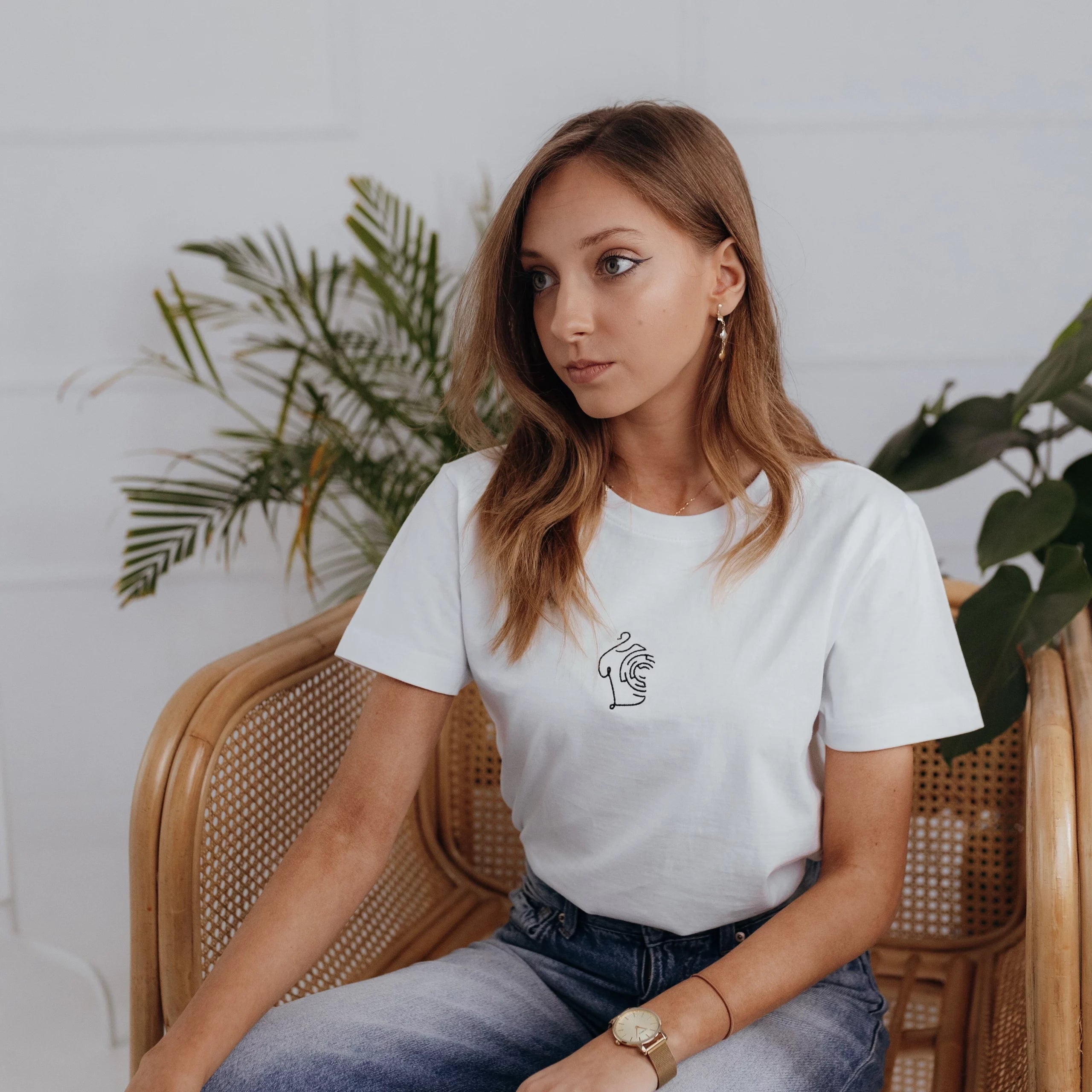 Kobieta w jeansach siedząca na bambusowym fotelu prezentuje klasyczny white minimaze t-shirt z wyhaftowanym, minimalistycznym wzorem na środku.