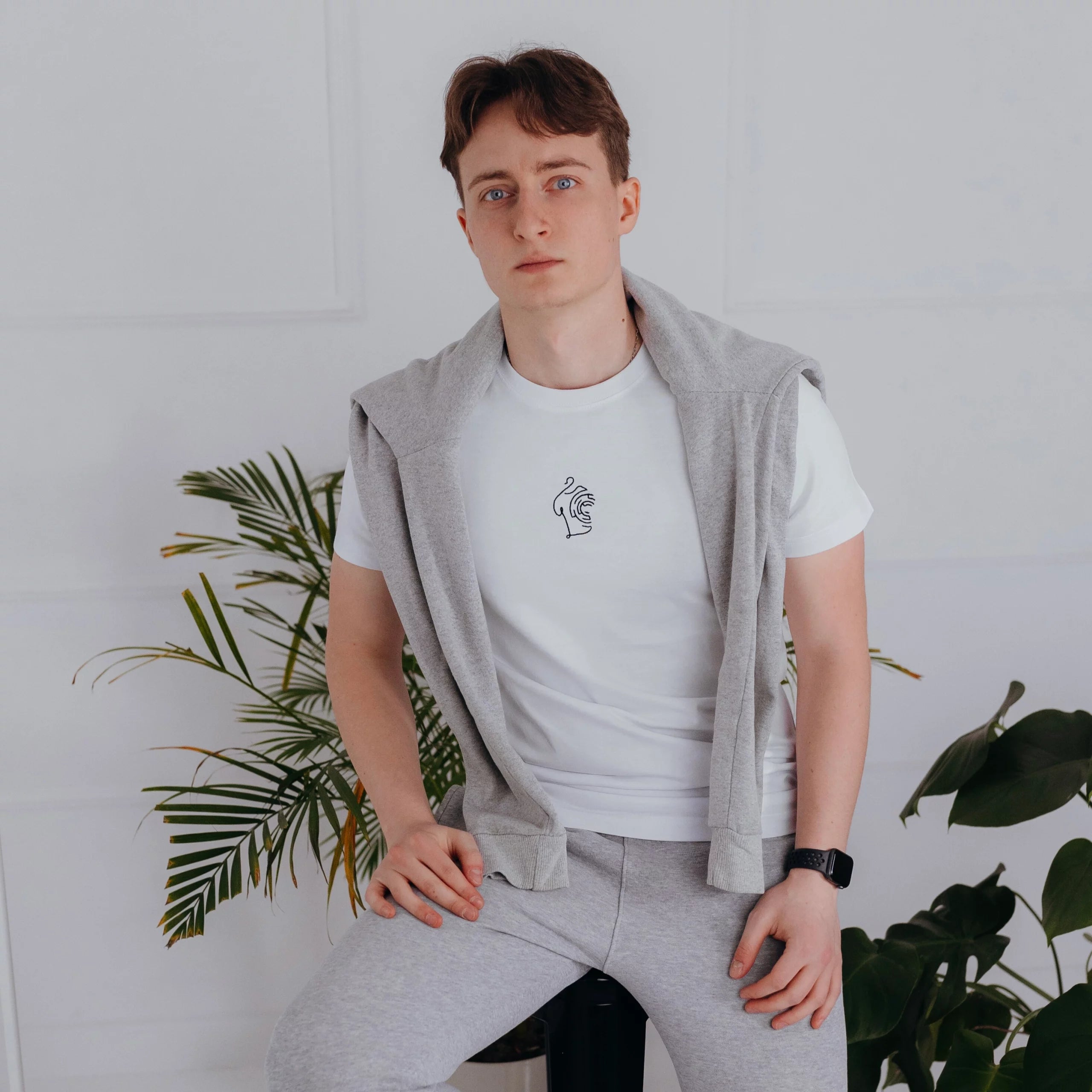 Mężczyzna w szarych spodniach dresowych prezentuje klasyczny white minimaze t-shirt z minimalistycznym haftem na środku oraz bluzą narzuconą na ramiona.