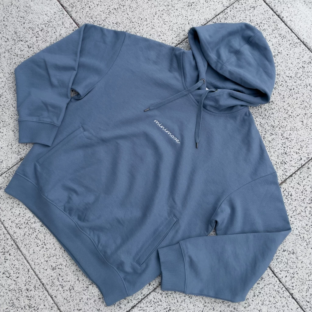 Niebieska bluza z kapturem marki minimaze, wykonana z miękkiego materiału, z minimalistycznym napisem 'minimaze' na klatce piersiowej. Bluza posiada kieszenie typu kangurka oraz regulowany kaptur, idealna na chłodniejsze dni.